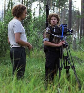Två personer står i en skog. En håller i ett filmstativ.