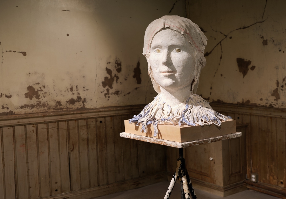 En skulptur av ett huvud täckt i vitt material, placerad på en träplattform som vilar på ett trebensstativ. I bakgrunden syns ett slitet rum med spruckna och flagnande väggar samt träpaneler.