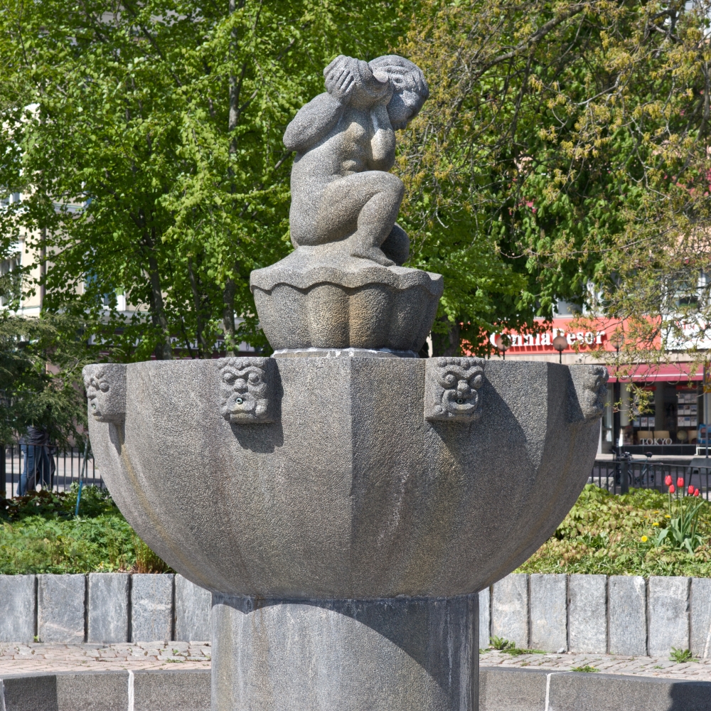 En stenfontän med en skulptur av en person som sitter ovanpå och täcker ansiktet med händerna. Fontänen har dekorativa element runt skålen, och i bakgrunden syns träd och byggnader.