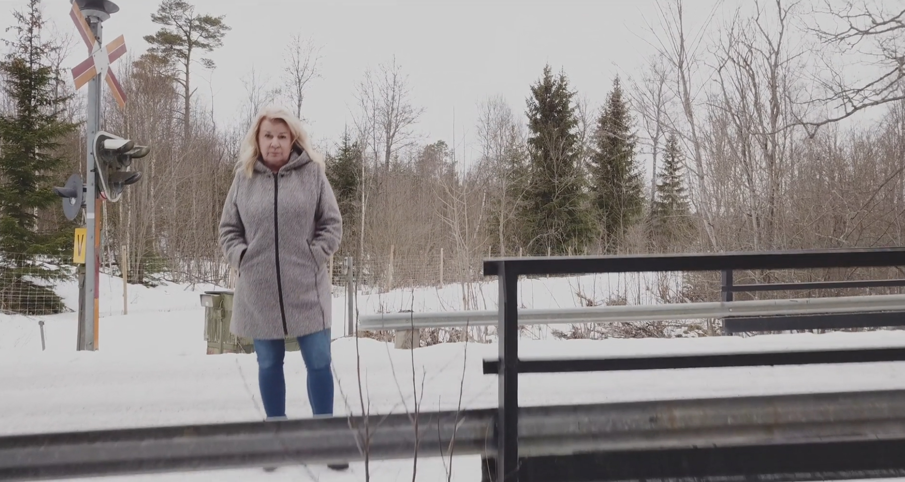 En kvinna står framför en skog. Hon kollar in i kameran och har händerna i fickan. Det är snö på marken.