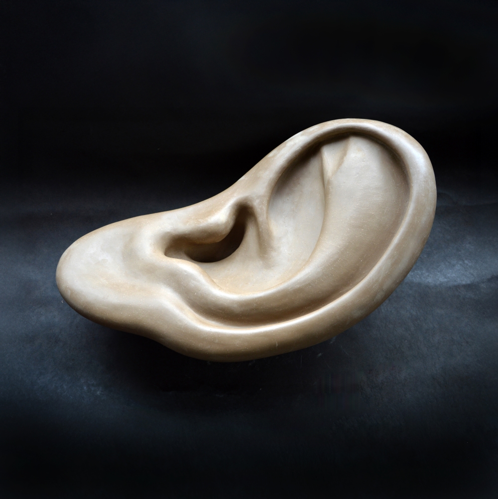 En skulptur av ett öra i ett slätt, beige material, avbildat i en abstrakt och överdriven form med mjuka kurvor och konturer, mot en mörk bakgrund.
