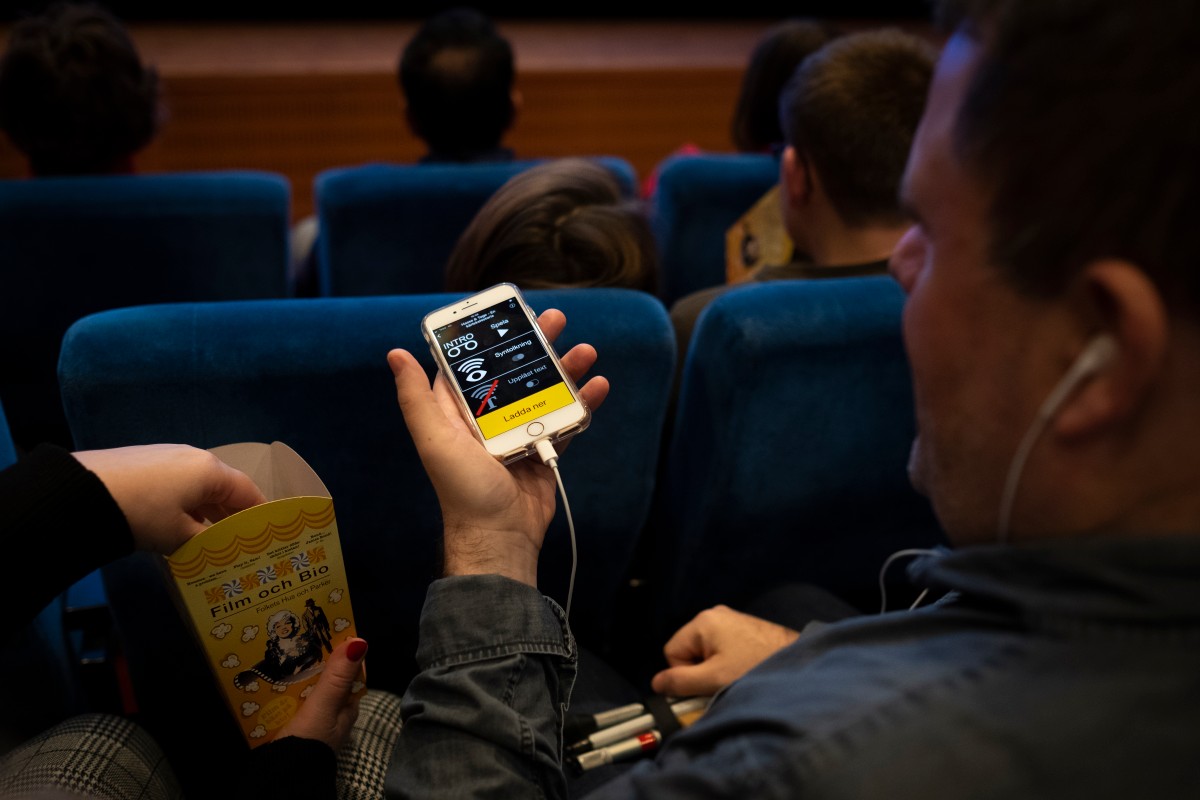 Bilden visar en person som sitter i en biosalong. Personen håller i en mobiltelefon med hörlurar inkopplade och verkar lyssna på något. Skärmen på telefonen visar en app för syntolkning. Personen har också en pappersförpackning med popcorn i andra handen. I bakgrunden syns andra personer som sitter i de blåa biosätena.