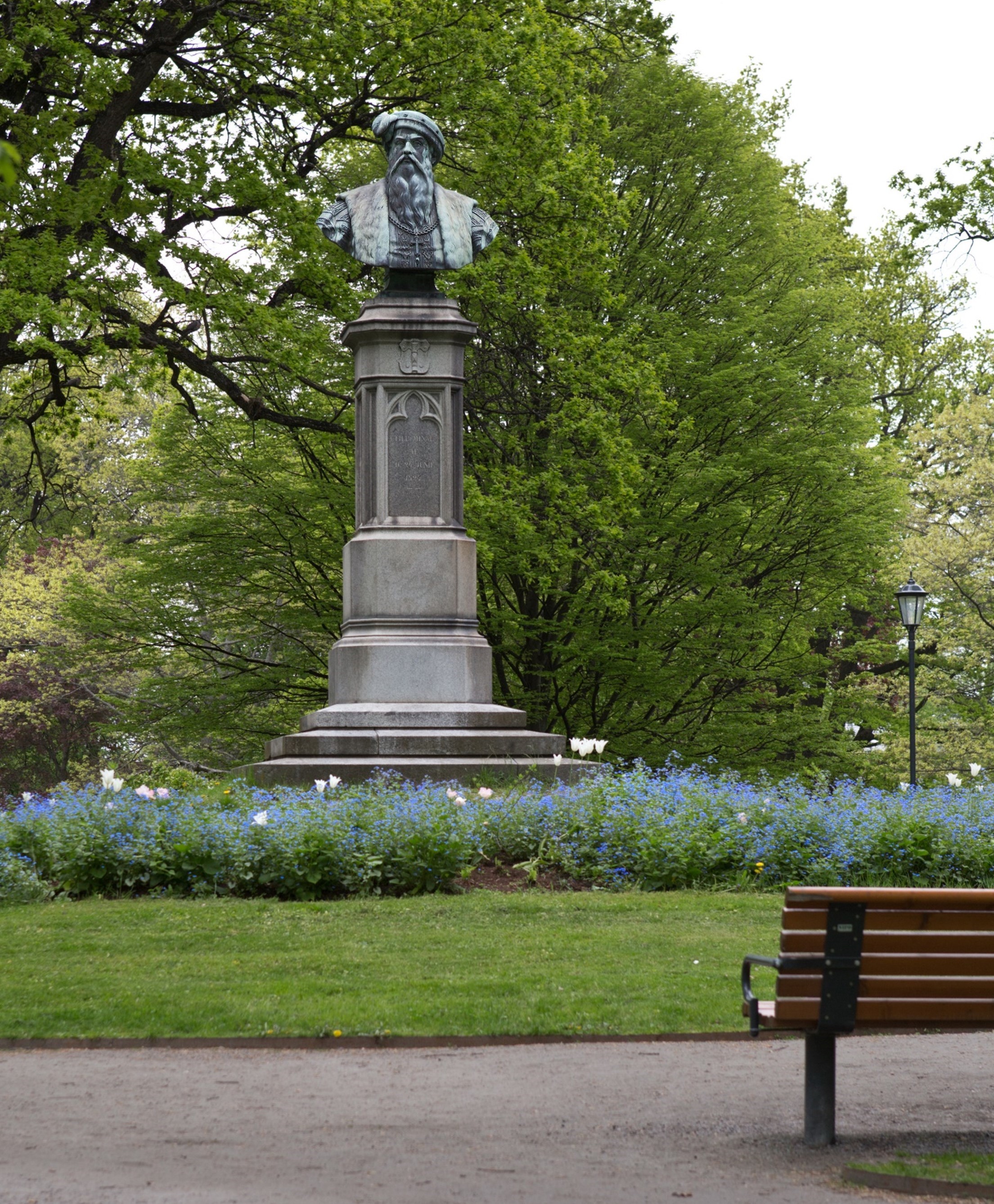 Bilden visar en staty i en park. Statyn föreställer en byst Gustav Vasa på en hög piedestal. Runt statyn finns det grönska och träd, och framför statyn finns en blomsterbädd med blå och vita blommor. Till höger i bilden syns en parkbänk.