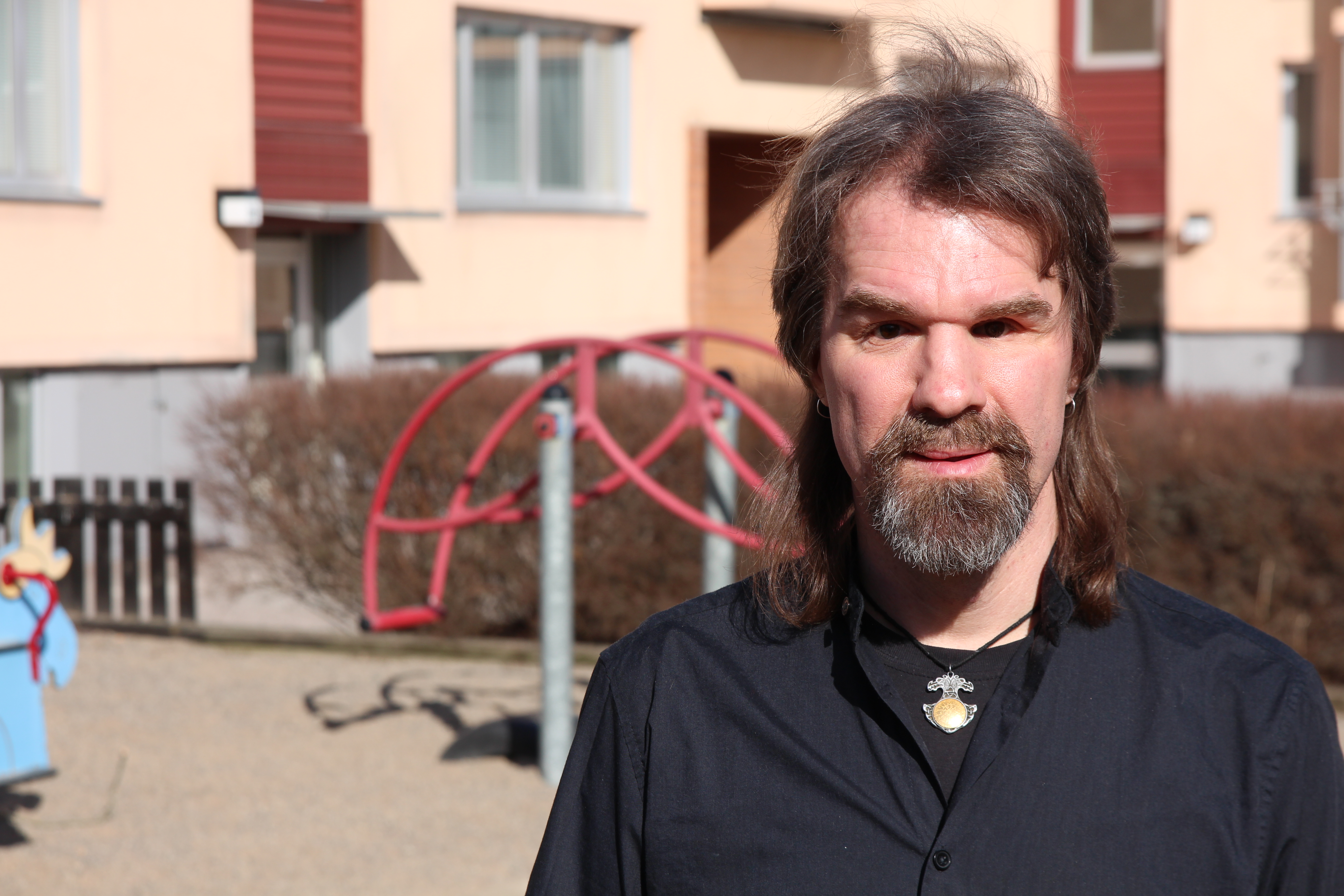 Fredrik Stockhaus Vänsterpartiet. Han är klädd i svart t-shirt och mörkblå skjorta, har ett halsband, långt mörkt hår och gråsprängt skägg. I bakgrunden ser man lekredskap i en lekpark i oskärpa.