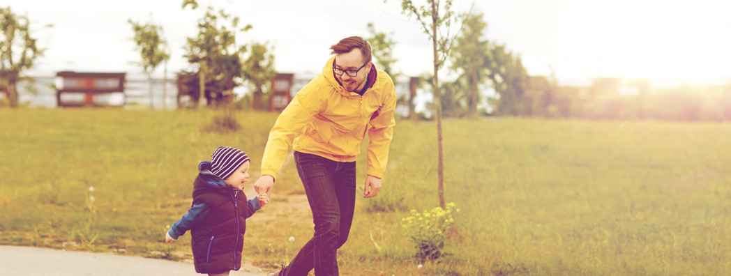 En man och ett yngre barn håller varandra glatt i handen. De är ute på promenad i höstsoligt väder.