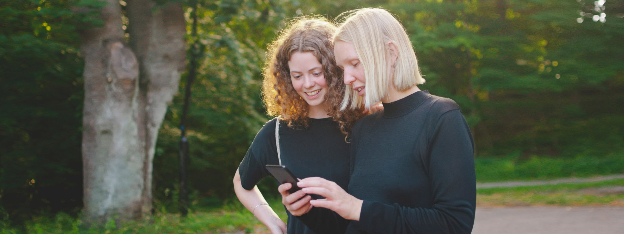 Två kvinnor står utomhus och tittar på en telefon.
