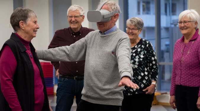 En äldre man testar VR-glasögon med vänner runtom som glatt kollar på.
