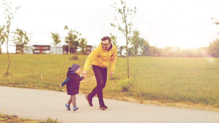 En man och ett yngre barn håller varandra glatt i handen. De är ute på promenad i höstsoligt väder.