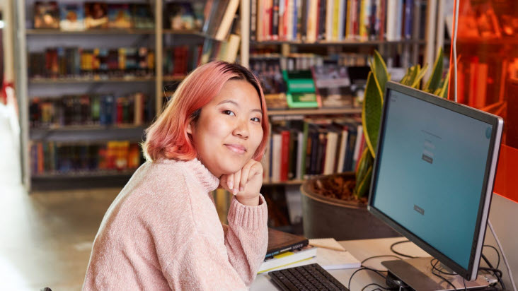 Ung kvinna med rosa hår sitter framför en dator i ett bibliotek