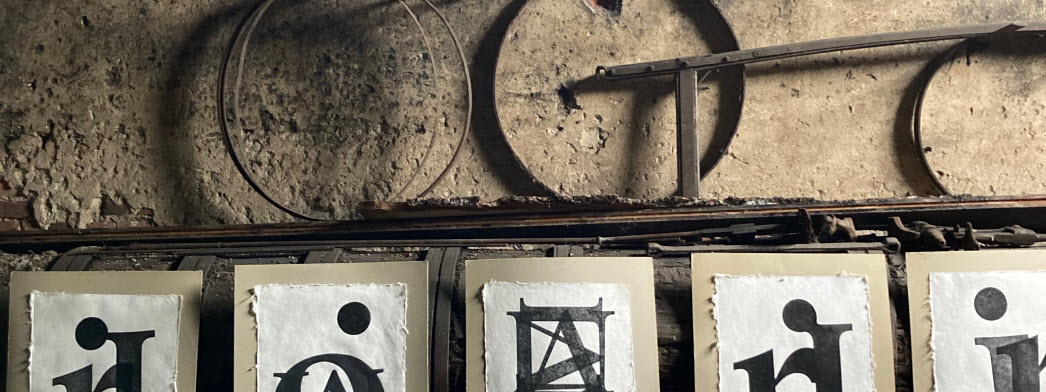Grafiska blad med abstrakta bilder genom tryck av stora bokstäver visas i industrihistorisk interiör jämte gamla verktyg som påminner om bokstäver.
