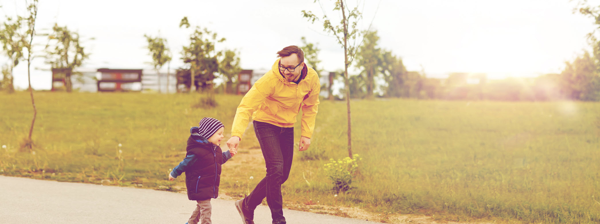 En man och ett yngre barn håller varandra glatt i handen. De är ute på promenad i höstsoligt väder.